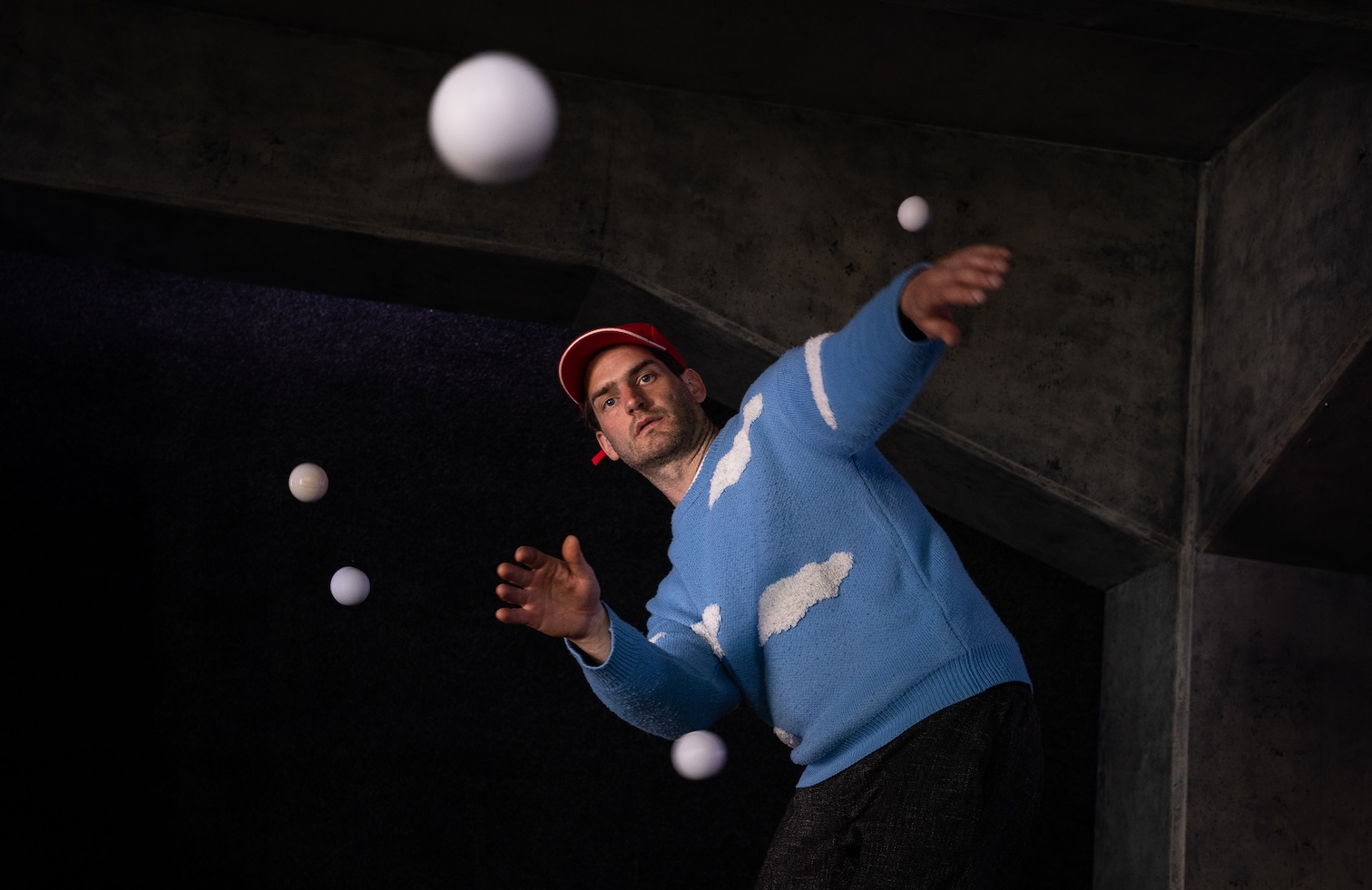 Mann mit roter Kappe und blaubem Pulli mit weißen Wolken. Um in herum fliegen viele weiße Bälle durch die Luft