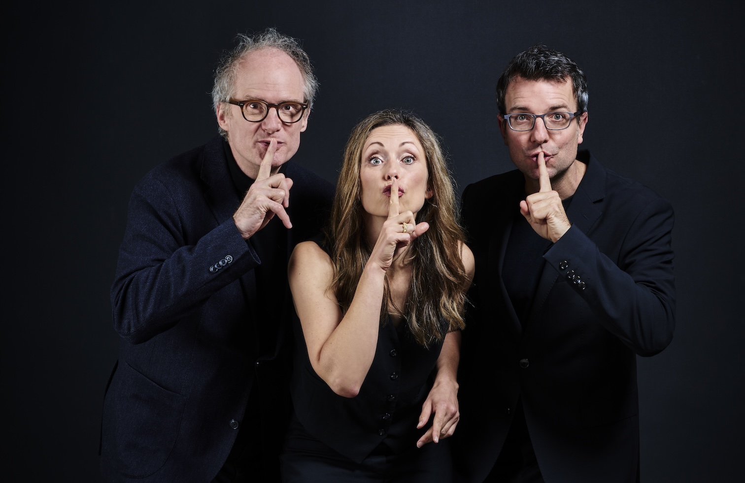 Eine Frau und zwei Männer in schwarzen Anzügen, die sich den Finger vor dem Mund legen als Zeichen zum leise sein